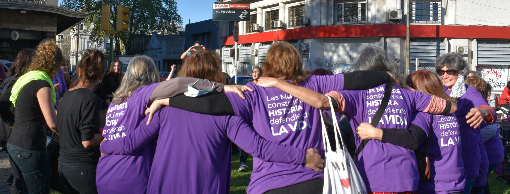 Fotografía. Un grupo de mujeres abrazandose en ronda. Tienen puesta una remera de color violeta con la frase "Las mujeres construimos historia defendiendo la vida".