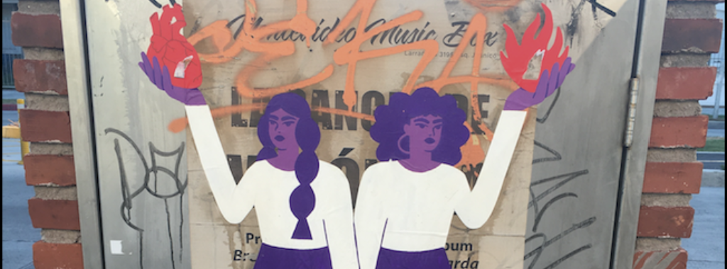 Fotografía. Una ilustración en una pared en la calle. Dos mujeres tomadas de la mano, con pelo y piel color tono violeta. Una de ellas sostiene un corazón y la otra una llama de fuego.