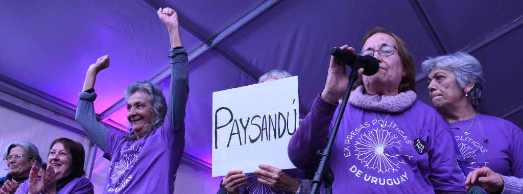 Fotografía. En un escenario mujeres del Colectivo Ex-presas políticas del Uruguay toman la palabra. Todas están usando una remera violeta con el logo del colectivo.Una de ellas tiene un cartel que dice "Paysandú", otra de ellas esta levantando los brazos.