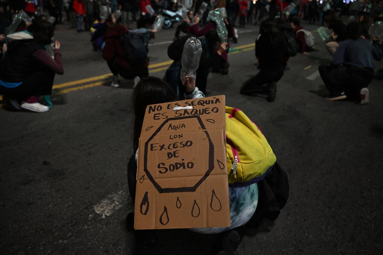 Fotografía. Una manifestación, las personas golpean el suelo con una botella vacía. Una mujer tiene un cartel que dice "No es sequía es saqueo", un dibujo de un octogano que adentro dice "Agua con exceso de sodio" y gotas de agua alrededor.