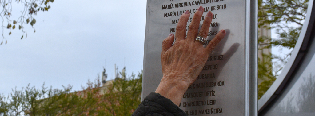 Fotografía. Una mujer pasa su mano en una estructura de metal dónde están los nombres de mujeres víctimas del terrorismo de Estado.