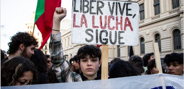 Fotografía. Exterior. Una manifestación en la calle. Una persona levanta el puño y sostiene un cartel que dice "Liber vive, la lucha sigue".